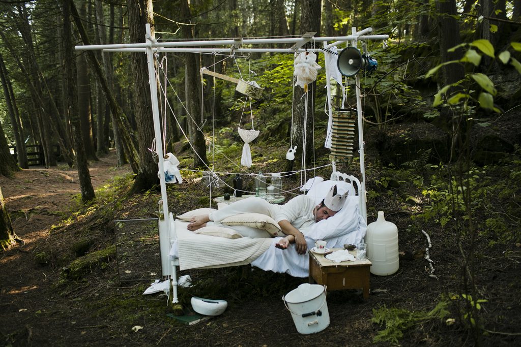 Dans cette image, une personne dort sur un lit en métal blanc au milieu d’une forêt, portant une couronne en papier. Le lit est recouvert de draps blancs, entouré de ficelles suspendues avec des décorations légères, créant une ambiance onirique. Une table de chevet en bois à côté contient des objets simples comme un bol et une bouteille. Le mélange du domestique et de la nature sauvage, avec une lumière douce, donne une impression de rêve.