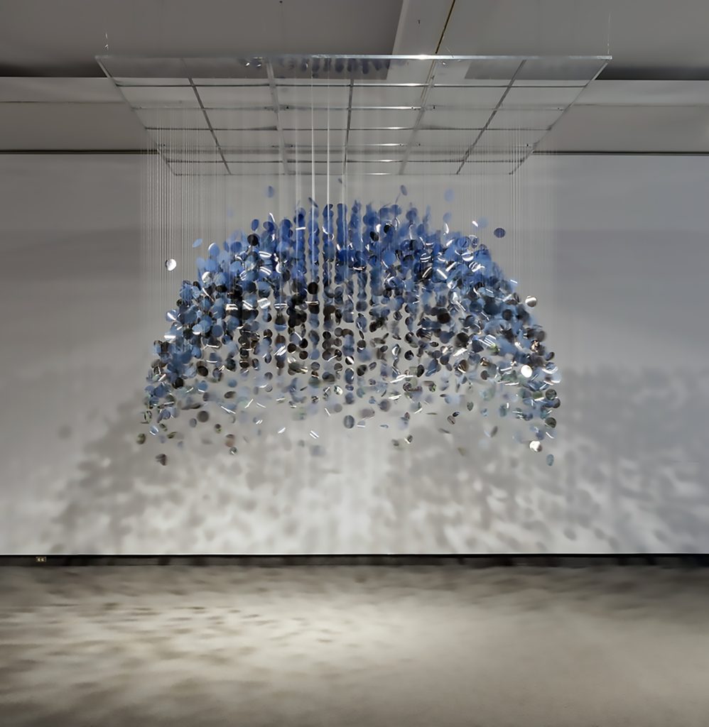 Cette image montre une installation artistique suspendue composée de nombreux petits objets circulaires, ressemblant à des disques ou des feuilles, accrochés à des fils fins. L'ensemble forme une structure en demi-sphère, avec une transition de couleurs allant du bleu en haut à des teintes plus sombres vers le bas. Les ombres des objets sont projetées sur le mur et le sol, créant un effet visuel supplémentaire.

La pièce semble être exposée dans une galerie d'art avec un éclairage qui accentue les détails et les ombres. L'effet général est éthéré et léger, avec une impression de mouvement ou de flottement.