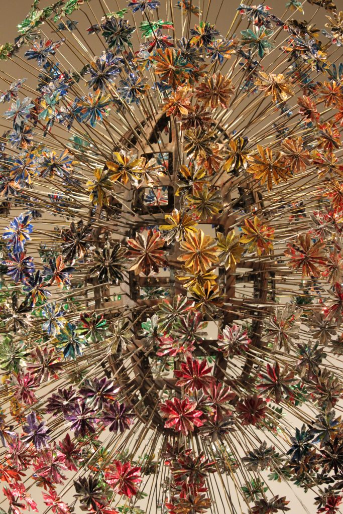 Cette photo montre une sculpture complexe composée de nombreux éléments en forme de fleurs, fabriqués à partir de matériaux brillants et colorés. Chaque "fleur" est reliée à une tige métallique qui converge vers un centre, créant une impression de dynamisme, comme si les fleurs émanaient du cœur de l’œuvre. Les couleurs vives – vert, bleu, rouge, jaune, et autres – contrastent avec les tiges métalliques, donnant à l'installation une apparence éclatante et vibrante. L'ensemble évoque un bouquet en explosion ou un feu d'artifice figé, avec un grand souci du détail et de la symétrie.