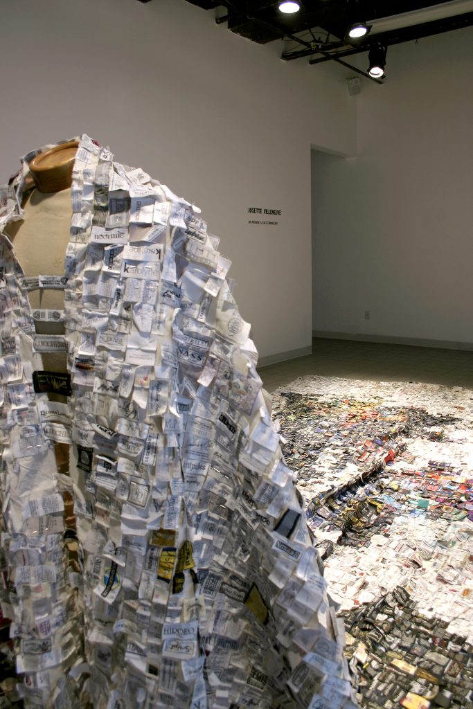 Cette photo montre une vue rapprochée d'une sculpture ou d'une installation artistique exposée dans une galerie. Au premier plan, on voit un mannequin recouvert d'une sorte de cape ou manteau, constitué de multiples petits morceaux de papier ou d'étiquettes, créant une texture dense et détaillée. Les morceaux semblent avoir été soigneusement disposés pour donner une apparence volumineuse à la cape. À l'arrière-plan, on aperçoit une continuation de l'œuvre qui s'étend au sol, probablement une partie de l'installation vue dans la première photo, faite de petits éléments similaires formant un motif ou une image. Le tout crée une impression de fragmentation et de détail méticuleux, reliant mode et art visuel.